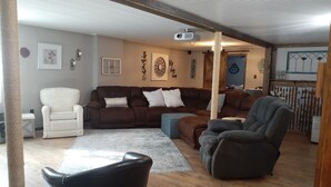 A living room with a brown sofa, a white armchair, and a grey recliner. There is a projector on the ceiling, a wooden floor, and a cat tree. - Beautiful house located 5 minutes drive to EAA AIRVENTUE (Oshkosh)