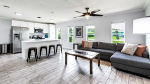 Eine moderne Küche mit einer zentralen Kochinsel, Barhocker, einem dunkelgrauen Sofa und einem Deckenventilator.
