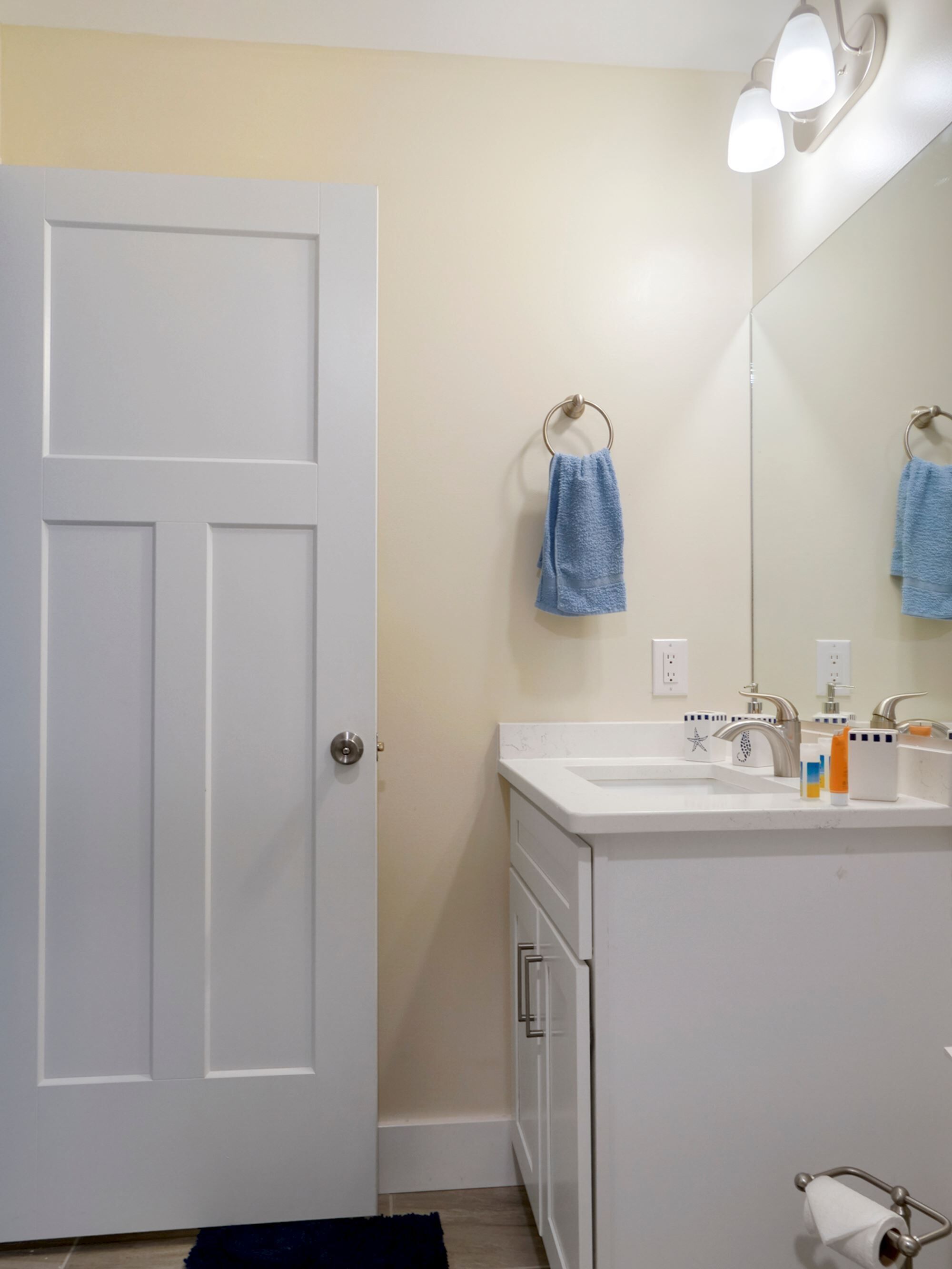 Un baño con una puerta blanca, un lavamanos con espejo y dos toallas colgadas en aros.