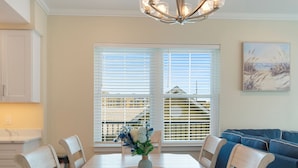 A dining area with a wooden table, chairs, and a chandelier. There are two windows with blinds, a painting on the wall, and a view of buildings outside. - Luxurious Bay Views steps Boardwalk (Ocean City)