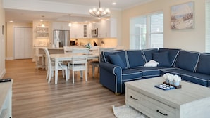 A modern living room with a blue sectional sofa, a wooden coffee table, and a dining area with white chairs and a kitchen in the background. - Luxurious Bay Views steps Boardwalk (Ocean City)