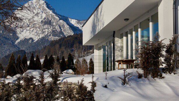 Un edifici modern amb grans finestres, un paisatge cobert de neu i muntanyes al fons.