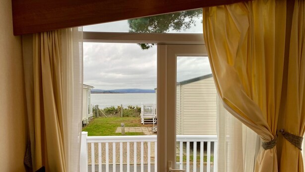 Blick aus einem Zimmer mit einer Schiebeglastür, die auf einen Balkon mit weißer Brüstung, eine Rasenfläche und ein Gewässer im Hintergrund führt.