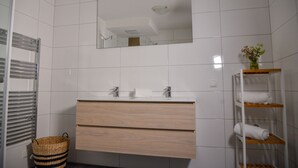 A modern bathroom with white tiles, a wooden vanity, a mirror, and a shelf with towels. - Grimming Lodge Goldrute (Tauplitz)