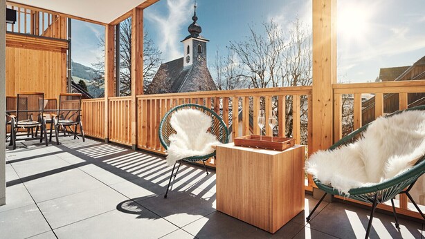 A wooden deck with two white furry chairs, a wooden table, and a view of a church with a bell tower. - Alm Lodge A2 - Tauplitz Lodges (Tauplitz)
