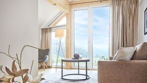 Uma sala de estar com um sofá, uma mesa redonda e uma cadeira. Há uma janela grande com cortinas, uma luminária e vista para o exterior.
