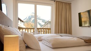 A bedroom with a bed, pillows, a lamp, a window with a view of mountains, and a TV on the wall. - Alm Lodge A3 - Tauplitz Lodges (Tauplitz)