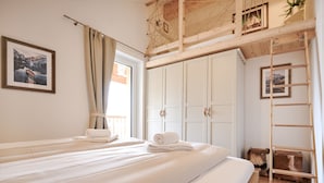 Une pièce avec un lit, une fenêtre avec des rideaux, une échelle en bois et un tableau encadré au mur.