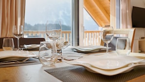 Une table à manger dressée avec des assiettes, des couverts et des verres à vin, donnant sur une terrasse en bois et avec un téléviseur visible au fond.