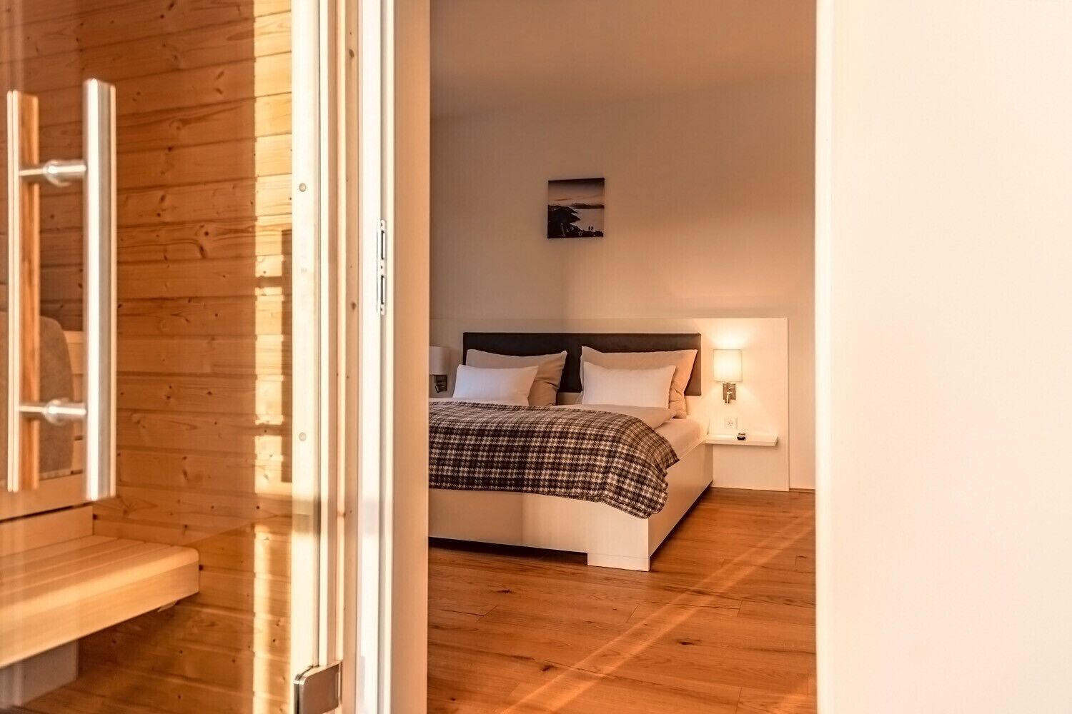 A bedroom with a wooden floor, a bed with a checkered blanket, a nightstand with a lamp, and a wall-mounted picture.