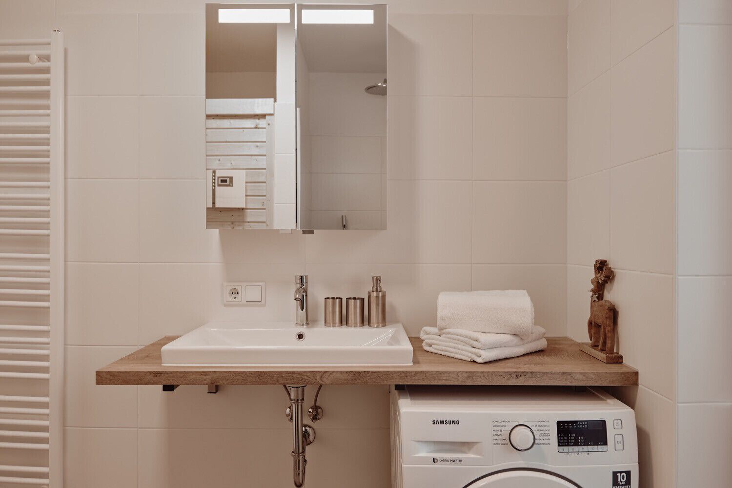 A bathroom with a white sink, a mirror above it, a wooden countertop, a stack of white towels, a washing machine, and a small statue on the right.