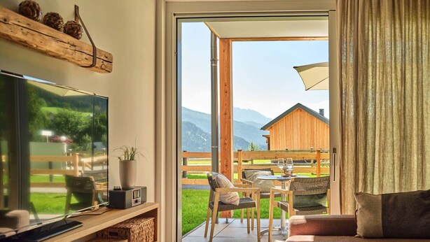 Ein modernes Schlafzimmer mit großem Fernseher, einer Holzwand mit Dekoration, einem gemütlichen Sofa und einer Glastür zum Außenbereich mit Tisch und Stühlen.