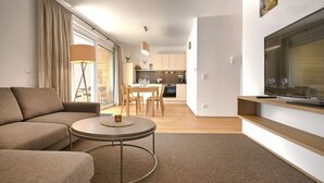 Un salon moderne avec un canapé, une table basse et un téléviseur posé sur un meuble en bois. On aperçoit une cuisine à l’arrière-plan, avec des meubles de rangement en bois et un coin repas.