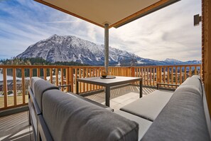 Un balcon doté d’une balustrade en bois, d’une table et de deux canapés gris. On aperçoit, au loin, une montagne dont les sommets sont enneigés.