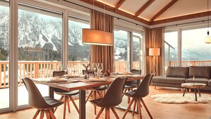 Un espace repas avec une table en bois, des chaises et une vue sur les montagnes, visible à travers de grandes fenêtres.