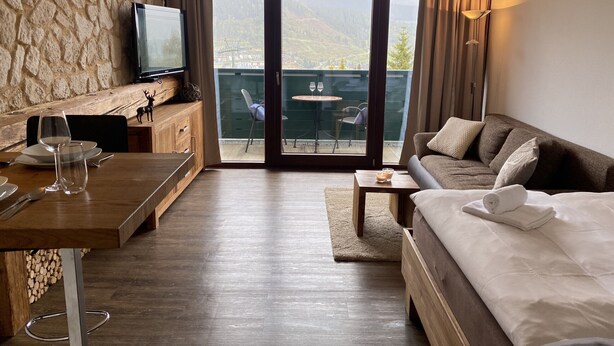 Een hotelkamer met een houten bureau, een televisie, een sofa en een balkon met uitzicht op bergen.