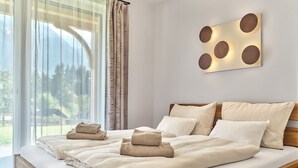 A modern bedroom with a wooden bed, white bedding, and a view of trees through sheer curtains. - Panorama Lodge Alpine Living 201 (Tauplitz)