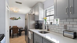 A modern kitchen with grey cabinets, a stainless steel sink, and a toaster. There is a dining table with chairs and a chandelier above. - Come Stay In This Charming & Cozy Home In The Heart Of Naples (Naples)