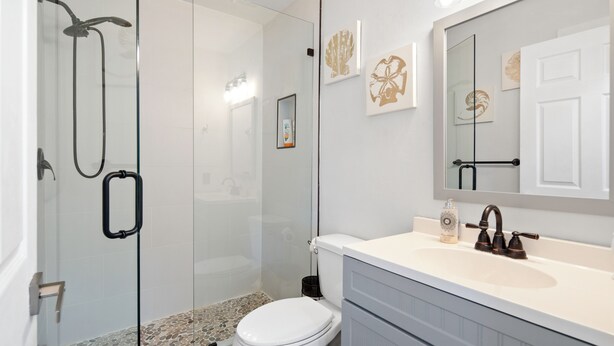 Une salle de bain avec une douche à parois de verre, des toilettes blanches, un meuble-lavabo avec un miroir et des œuvres d’art décoratives au mur.