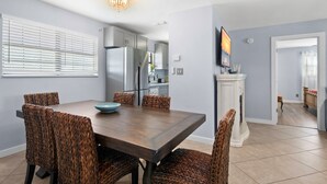 A dining area with a wooden table and woven chairs, a kitchen in the background, and a fireplace with a TV above it. - Come Stay In This Charming & Cozy Home In The Heart Of Naples (Naples)