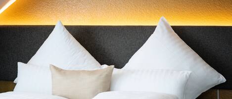 Una cama bien tendida con almohadas blancas y beige, cabecera oscura y una cálida fuente de luz arriba.