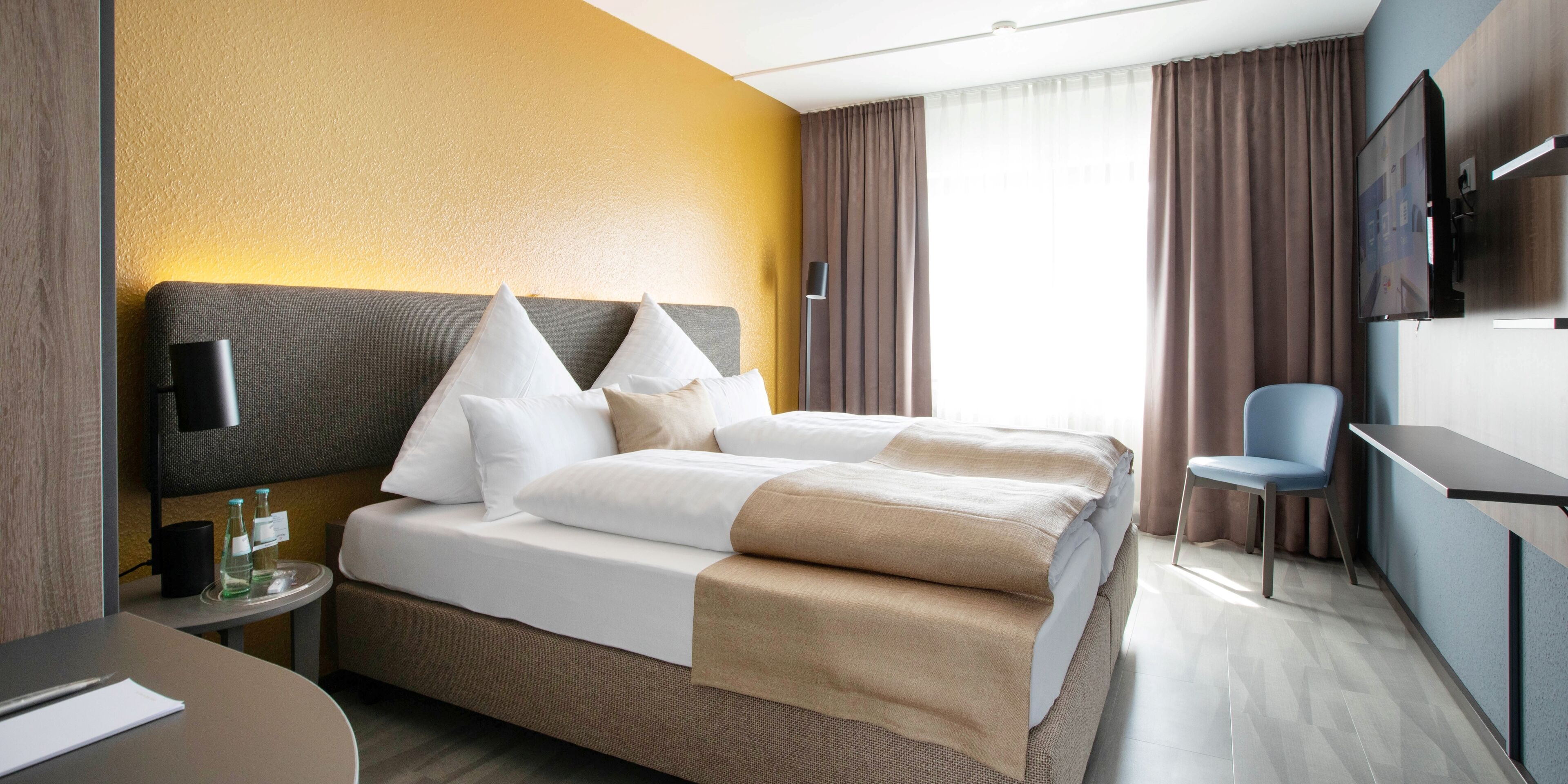 Habitación de hotel con cama, mesita de noche, silla y televisión montada en la pared.
