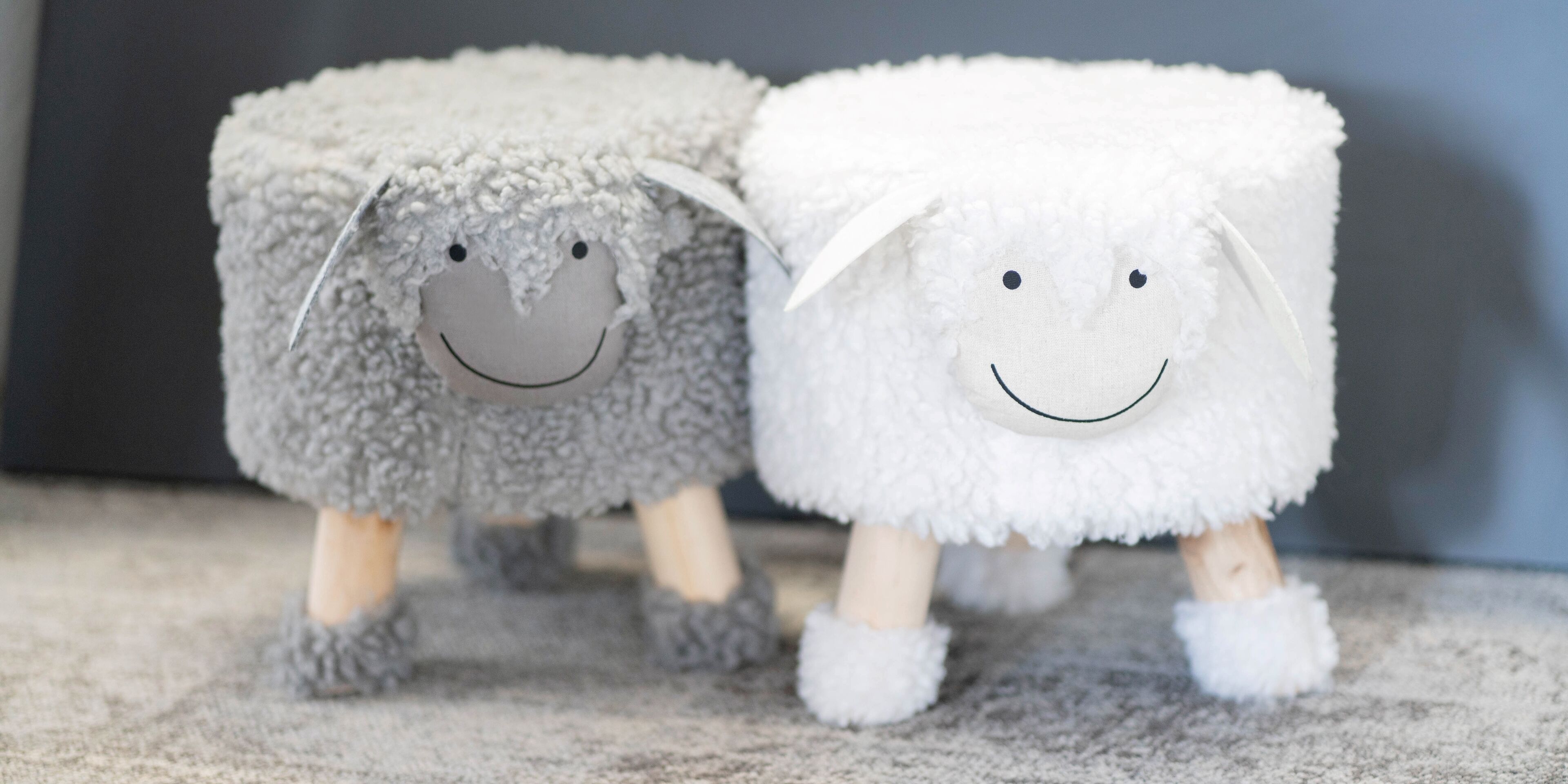 Two plush sheep figures with smiling faces and wooden legs standing on a carpet.
