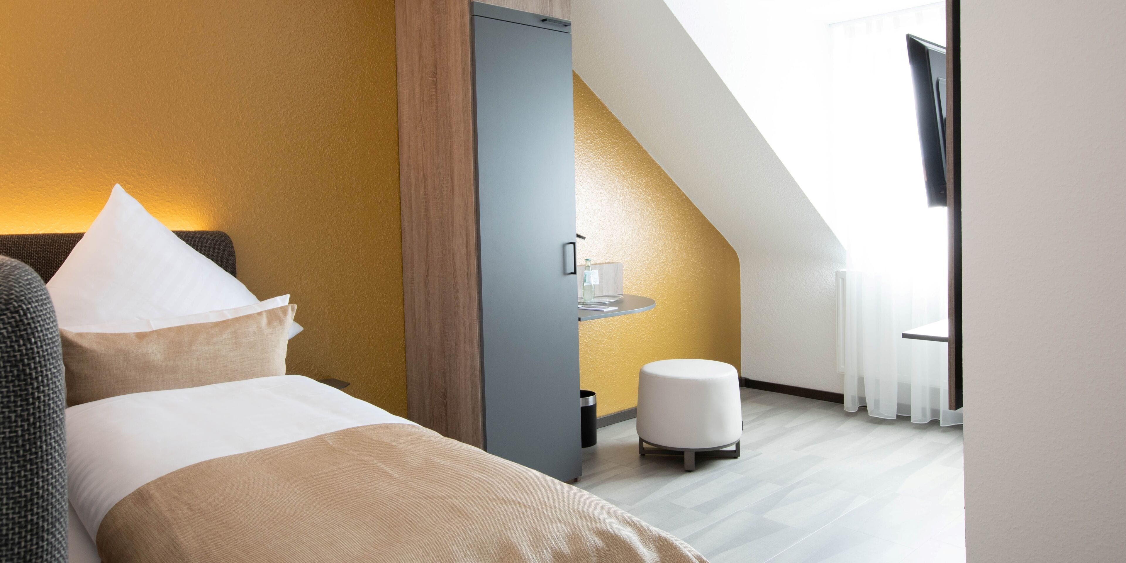 A modern hotel room with a bed, a bedside table, a small stool, and a window with curtains.