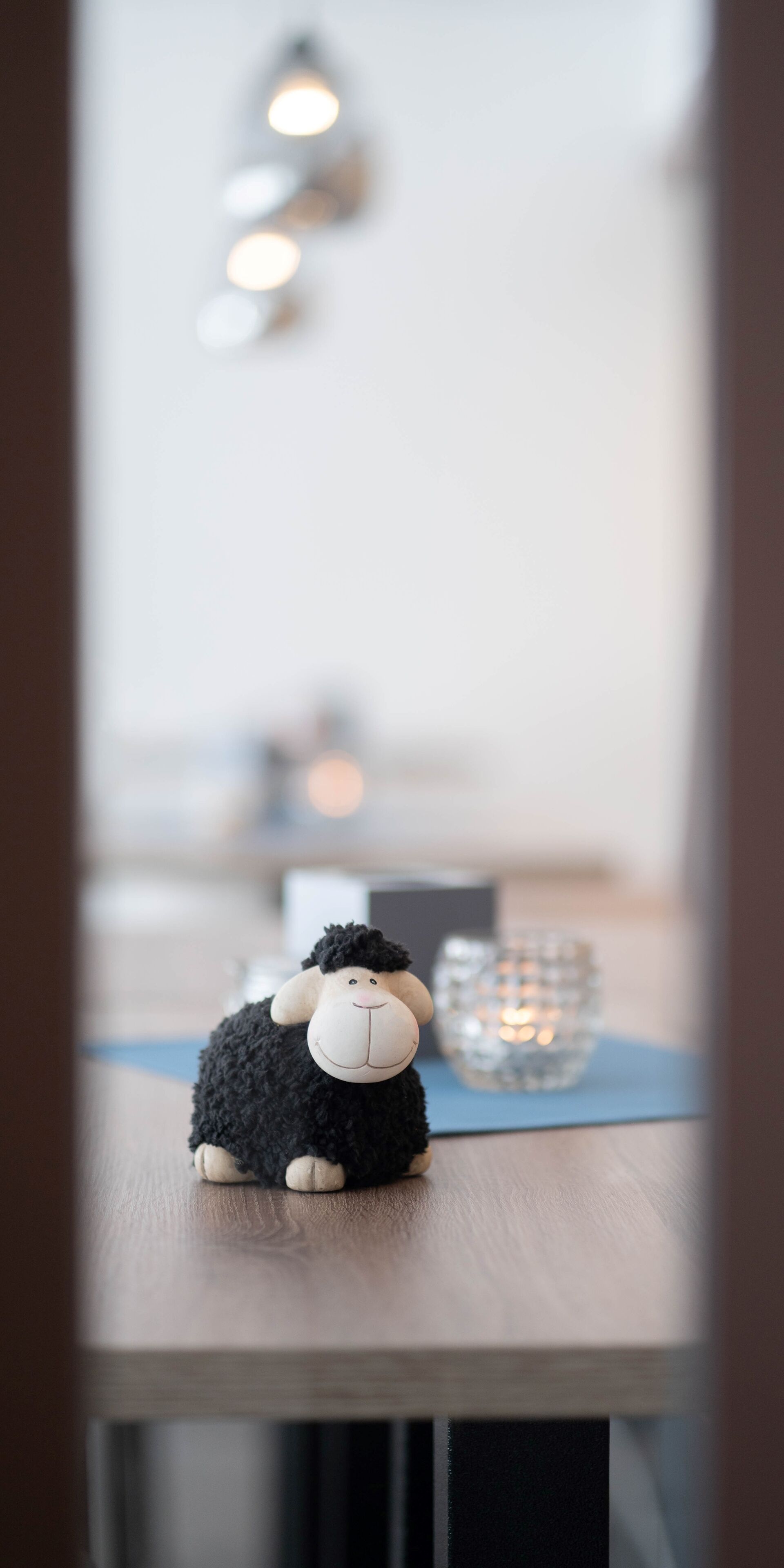 Un juguete de oveja negra mullida sobre una mesa de madera, con un candelabro de vidrio al fondo.