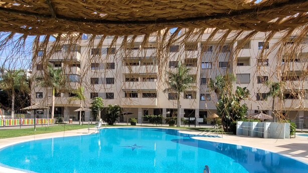 Une piscine à l’eau d’un bleu limpide, entourée d’une terrasse carrelée. Au fond, on aperçoit un immeuble à plusieurs étages doté de balcons et une structure à toit de chaume au-dessus de la piscine.