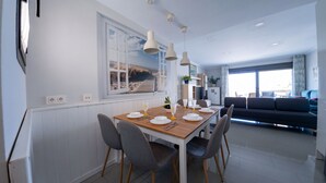 Un coin repas avec une table en bois dressée pour quatre personnes, des chaises grises, des luminaires suspendus, une grande fenêtre donnant sur la plage et, au fond, un canapé de couleur sombre.