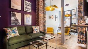 A modern living room with a green sofa, colorful cushions, a glass coffee table, and a yellow armchair. There are framed pictures on the wall, a brick accent wall, and a dining area with yellow chairs. - Appart indépendant, 2chambres!Parc des expositions (Paris)