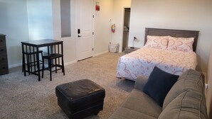 A bedroom with a bed, a sofa, a small table with two chairs, a black ottoman, and a door with an exit sign. - CASITA NEAR ZION (Hurricane)