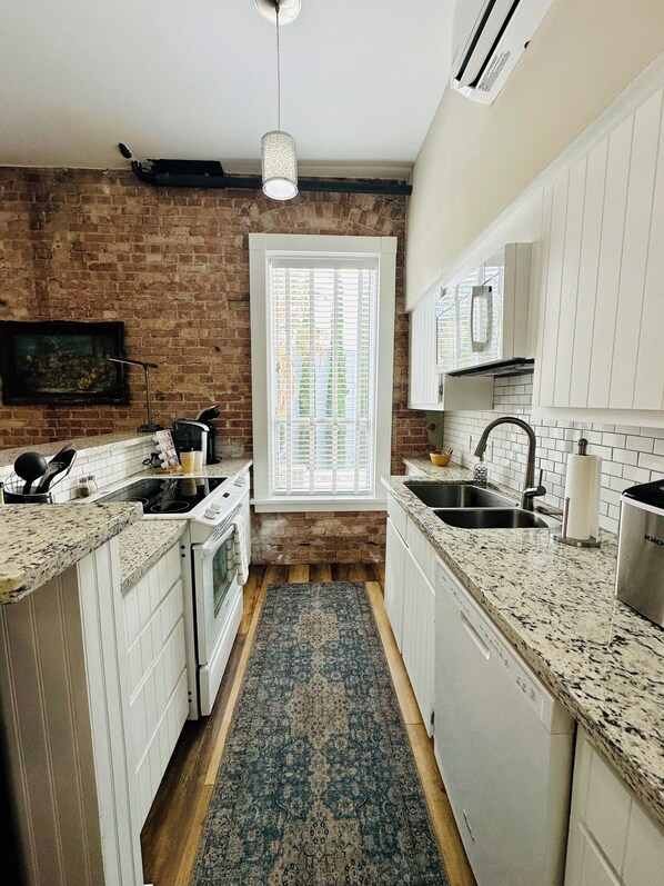A kitchen with white cabinets, a brick wall, a window with blinds, and a patterned rug.
