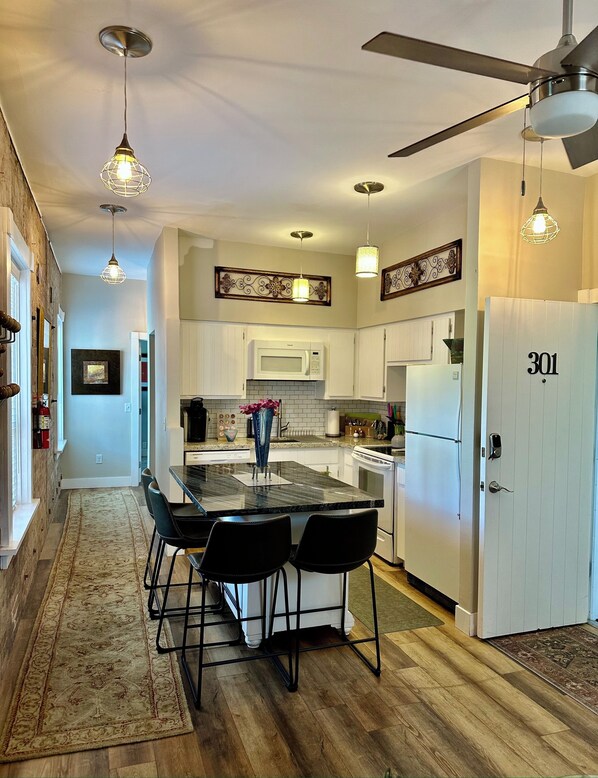 A modern kitchen with white cabinets, a central island with bar stools, a microwave, and a refrigerator. - Bisbee Brownstone Suites (Bisbee)
