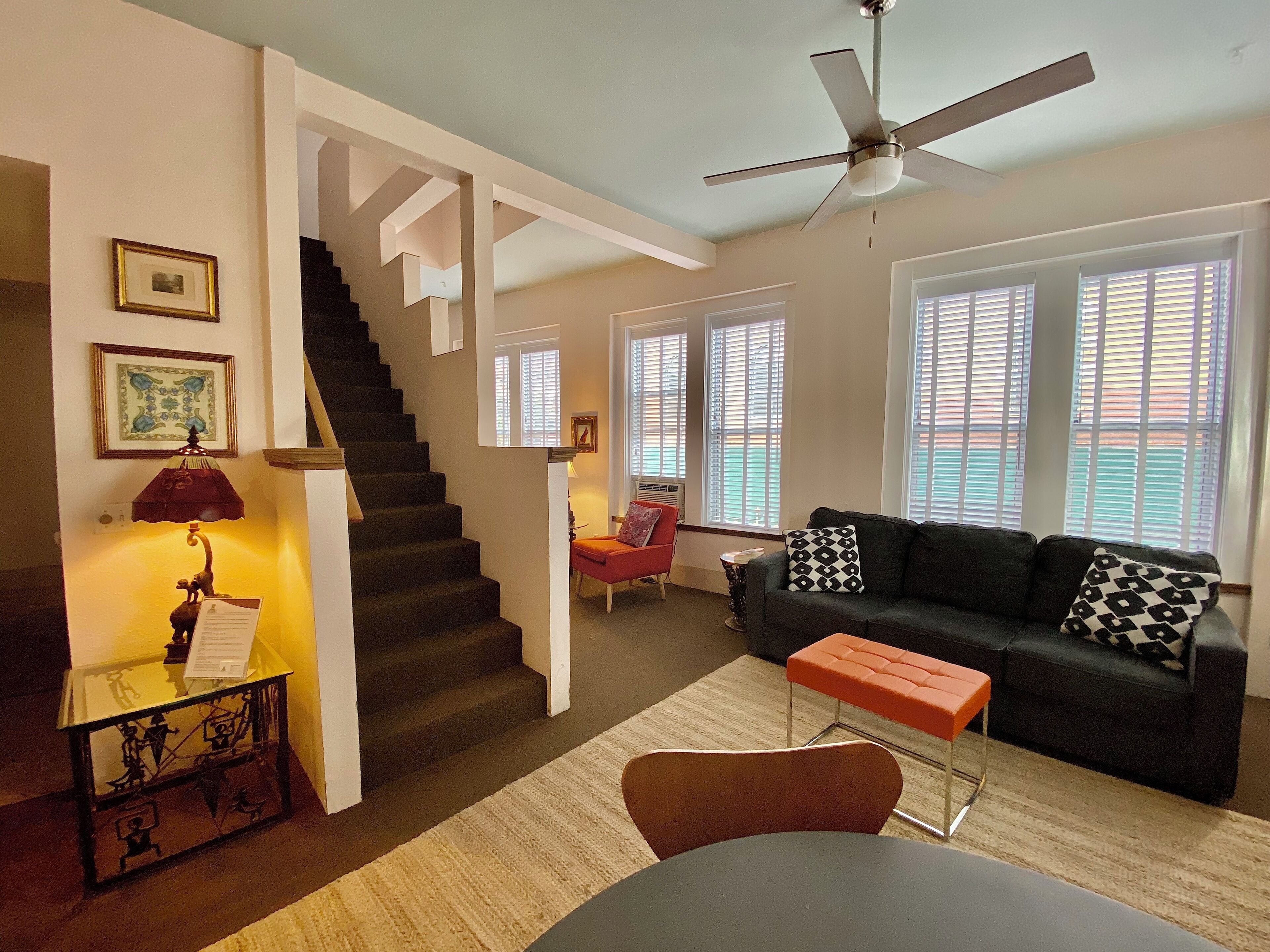 A living room with a staircase, a ceiling fan, a sofa with patterned cushions, and a table with a lamp.
