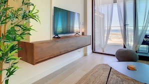 Ein modernes Wohnzimmer mit einem flachbildfernseher an einer Holzkonsole, einem großen Fenster mit transparenten Vorhängen und Blick ins Freie.