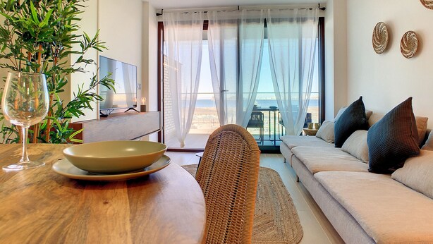 Ein modernes Wohnzimmer mit einer Couch, einem Esstisch mit Schüssel und Weinglas, einem Fernseher und Meerblick durch transparente Vorhänge.