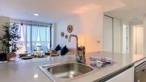 Eine moderne Küche mit Spüle aus Edelstahl, einem Essbereich mit Tisch für zwei Personen und Blick ins Freie durch ein Fenster mit leichtem Vorhang.