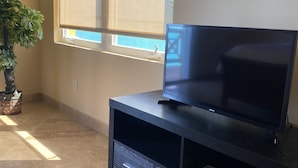 A living room with a flat-screen TV on a dark wooden entertainment unit, a potted plant, and a large window with blinds. - The Four Breezes With Pool and View (providenciales)