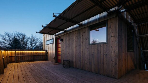 Una terraza de madera con una puerta roja y una ventana, bajo un techo de metal.