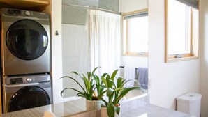 Un baño pequeño con lavadora, ducha con cortina, una ventana pequeña y una planta en maceta sobre el mostrador.