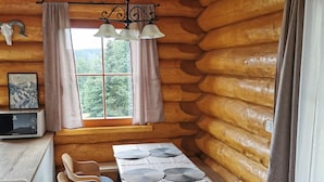 A cozy log cabin interior with a dining table, chairs, a small kitchen area, and a window with curtains.