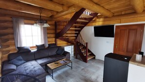 A cozy log cabin interior with a sofa, a wooden table, a staircase, and a TV mounted on the wall.