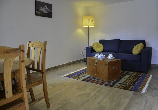 A living room with a wooden dining table, a blue sofa with yellow cushions, a lamp, and a small wooden chest with teacups. - Nepal in Queyras (Queyras)