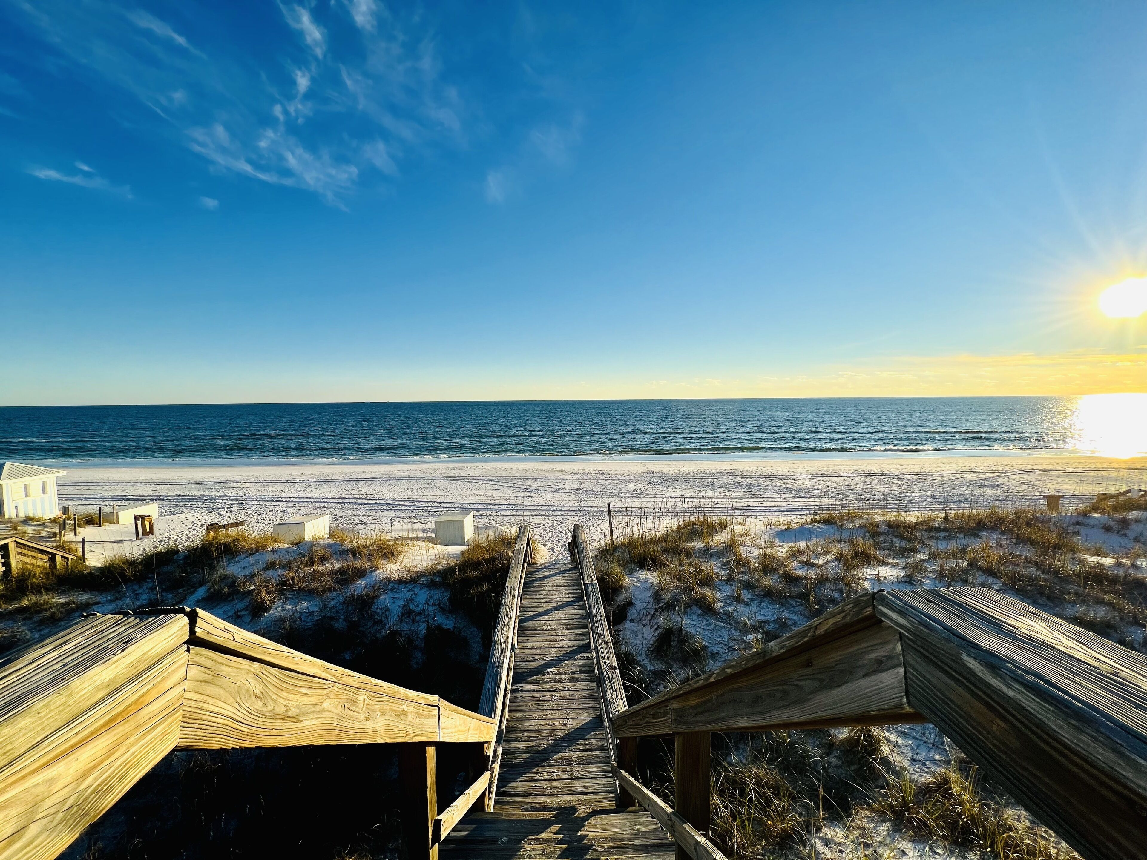 A wooden boardwalk leading to a beach with a clear sky and the sun setting in the background.