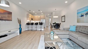 Uma sala de estar moderna com sofá, mesa de centro e um aparador para TV. Ao fundo, há uma área de cozinha com banquetas e uma mesa de jantar.