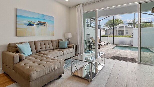 Uma sala de estar moderna com sofá de canto, mesa de centro de vidro e vista para uma piscina e área externa com mobiliário.