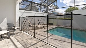 Área de piscina com estrutura de vidro, cadeiras de descanso e vista clara do céu e das árvores.