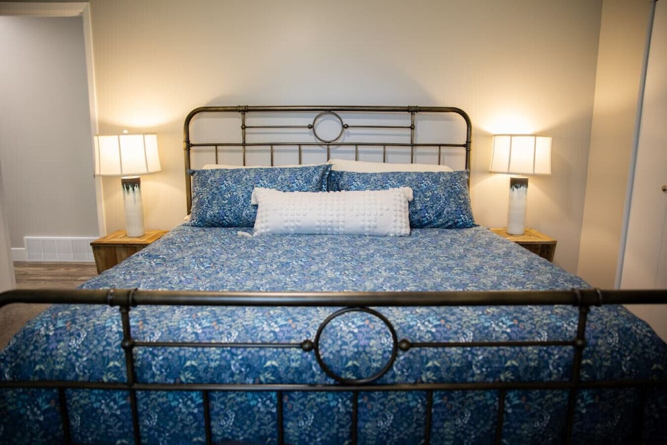 Ein Bett mit Metallgestell und einer zudeckenden Bettwäsche mit Blumenmuster. Zwei Nachttische mit Lampen stehen daneben.
